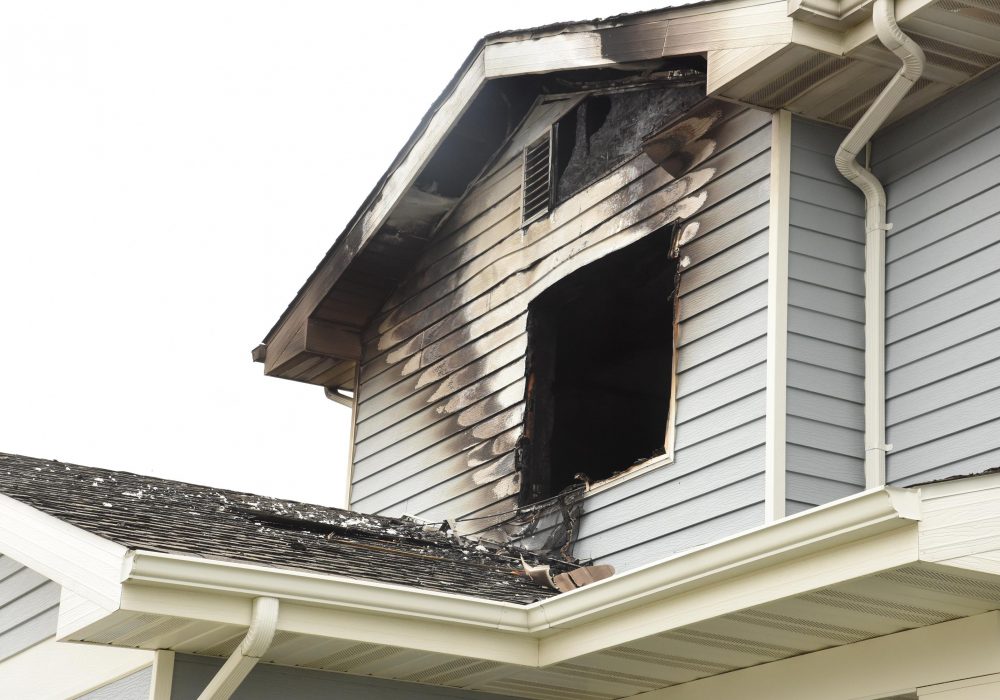 A picture of the exterior of a blue house. Parts of the roof, siding, and window are blackened from fire damage.