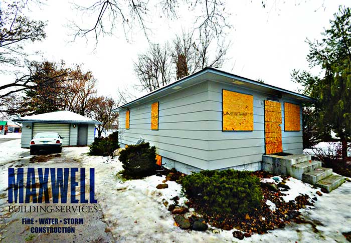 an image of a small, blue home that has been boarded up after fire damage. There is snow and ice on the ground. Maxwell Building Services's blue logo is in the bottom left corner along with the words "Fire, Water, Storm Construction"